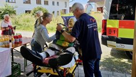 Der Gast der Herzenswunsch-Fahrt liegt lächelnd auf einer fahrbaren Komfortliege, daneben stehen ein Mann mit Malteser-Herzenswunsch-T-Shirt und eine Frau. 