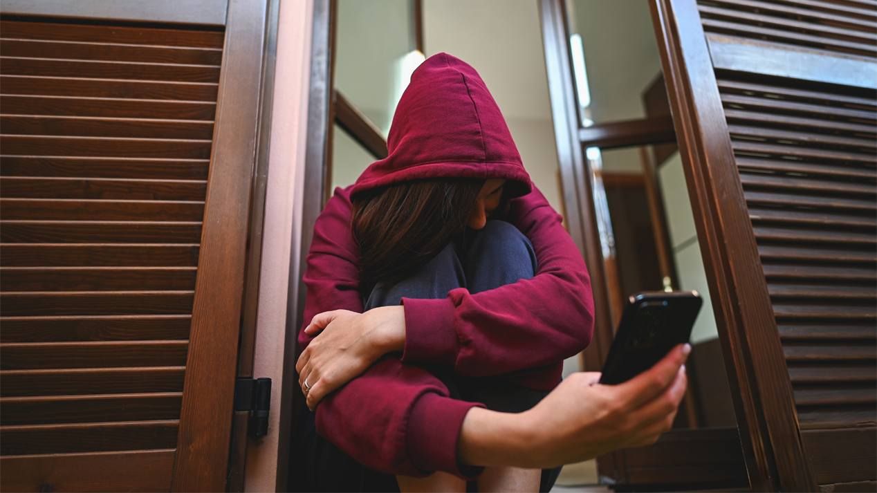 Eine junge Frau mit einer Kapuze über dem Kopf sietzt und schaut auf ein Smartphone 
