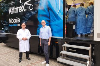 Zwei Männer stehen vor einem mobilen Trainingslabor, in dem Personen in OP-Kleidung arbeiten.