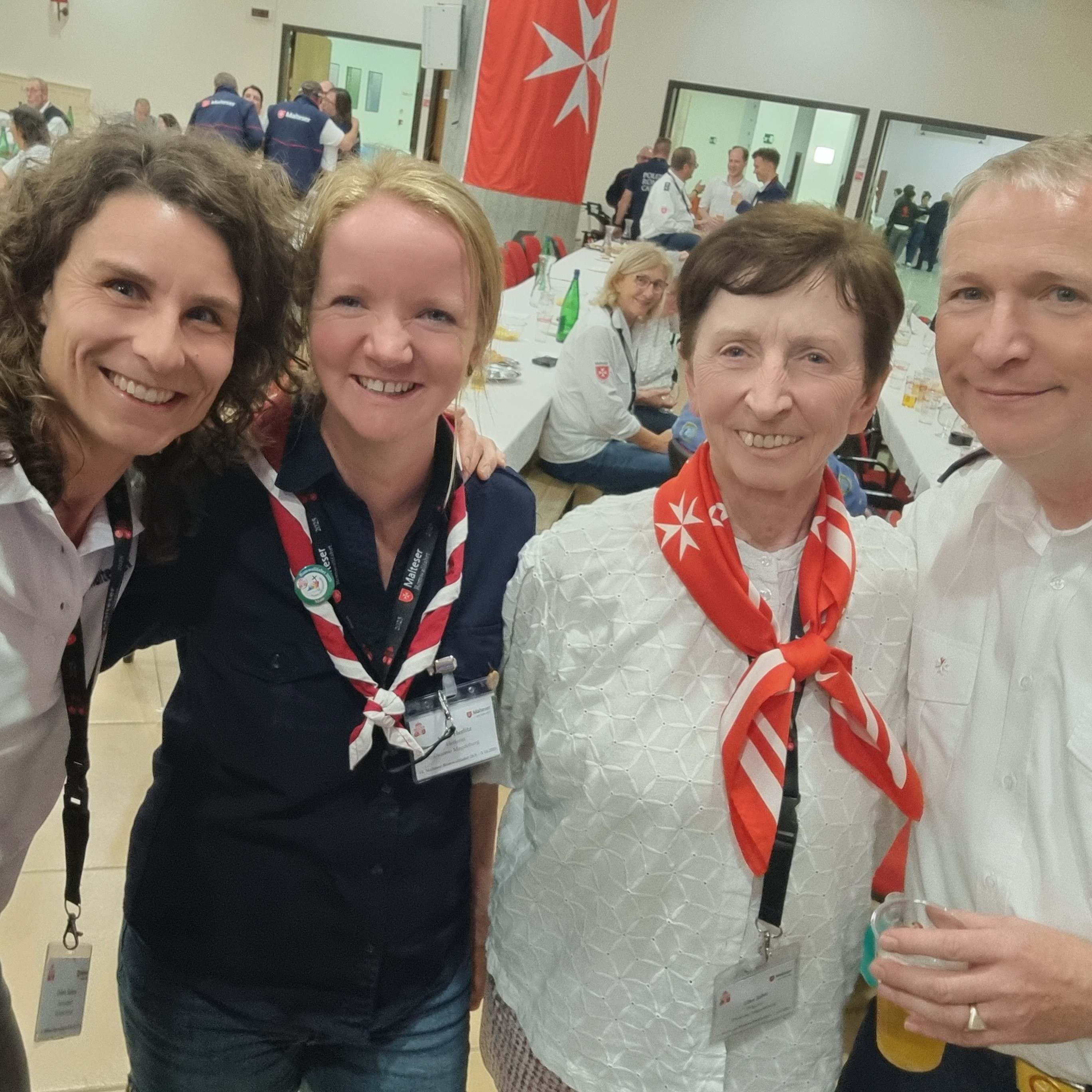 Vier Personen in weißer und schwarzer Kleidung mit roten Halstüchern in einem hellen Raum mit rotem Malteser Banner an der Wand, Veranstaltung Festa Romwallfahrt.