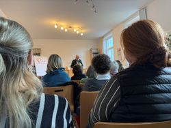 Mehrere Personen sitzen auf Stühlen in einem hellen Raum und hören einer vortragenden Person zu, die vorne an einem Tisch steht und aus einem Buch liest. Im Hintergrund befinden sich Regale mit Büchern und Ordnern sowie ein Flipchart mit einem Herz‑Motiv. Von der Decke hängen mehrere warm leuchtende Spotlampen, und durch die großen Fenster fällt Tageslicht in den Raum.