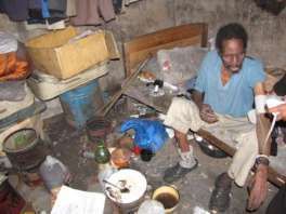 A woman sits in a simple hut among household items.