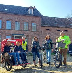 2 Frauen und ein Hund sitzen in einer Rikscha. 4 weitere Personen mit Fahrrad und ein Hund mit Fahrradkorb stehen rechts daneben. (Foto: Malteser Westpfalz)