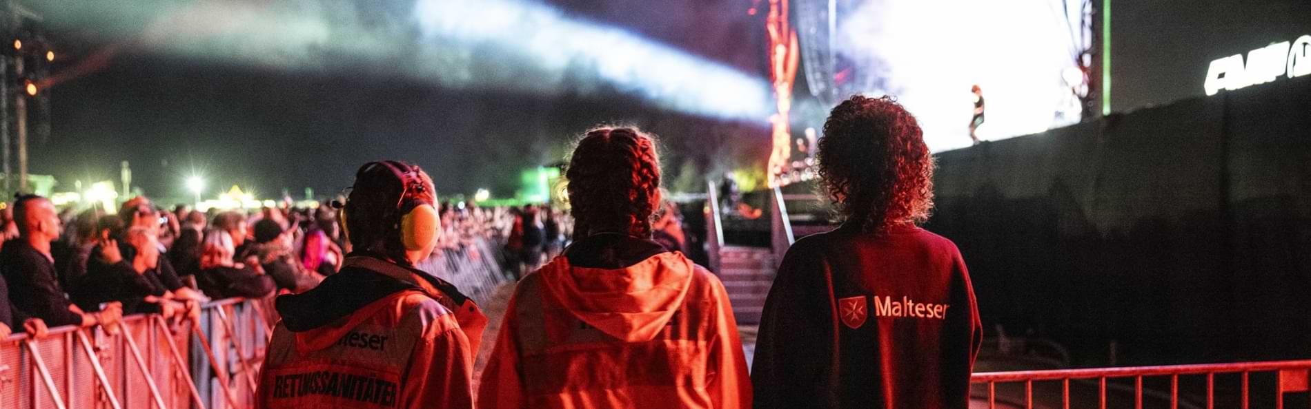 Rückansicht von drei jungen Frauen in Einsatzkleidung des Malteser Hilfsdienst im Bühnengraben eines Festivals. 