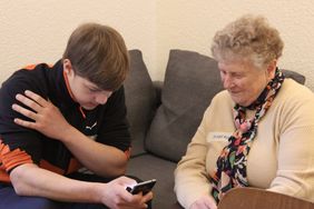 Ein junger Mann sitzt auf einer grauen Couch und bedient ein Handydisplay. Eine ältere Dame sitzt daneben und schaut neugierig und lächelt. 