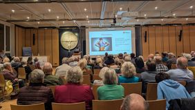 Viele Personen sitzen mit dem Rücken zur Kamera in einem Raum und hören sich einen Vortrag an.