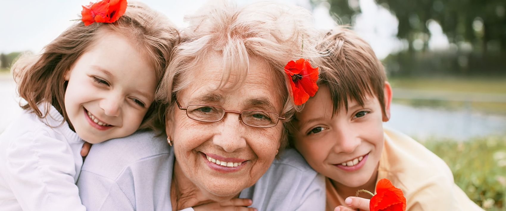 Eine ältere Frau lacht mit zwei Kindern in die Kamera. Sie haben Blumen im Haar.