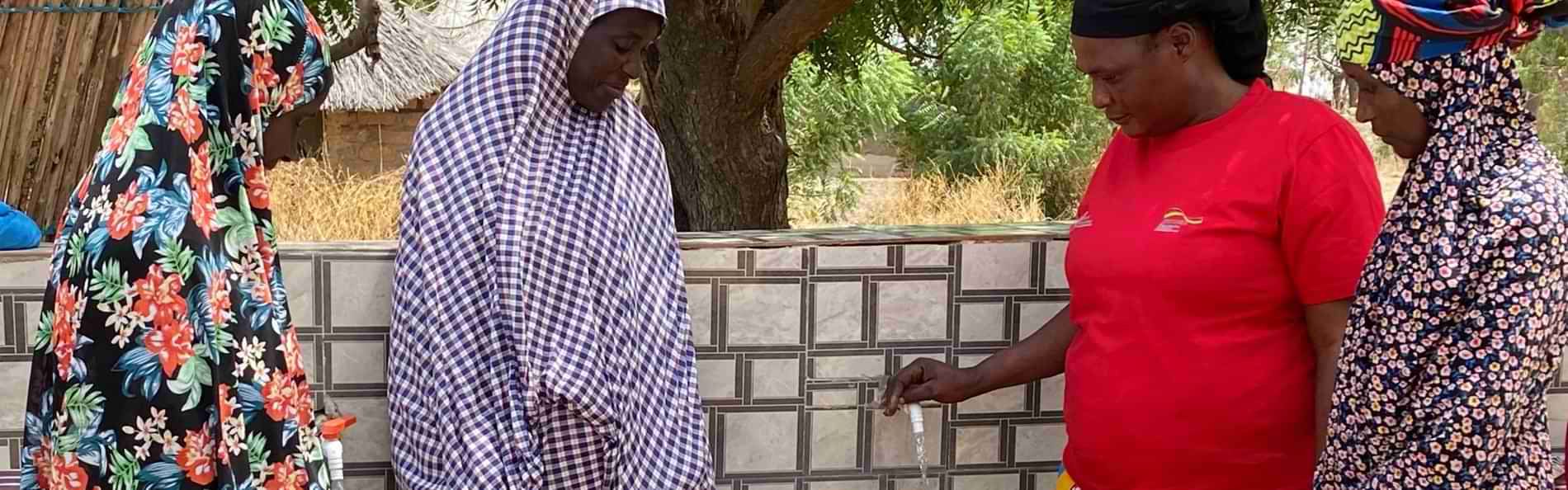 Eine Mitarbeiterin von Malteser International und drei weitere Frauen stehen im Freien um einen Wasserhahn und füllen Kunststoffeimer mit Wasser.