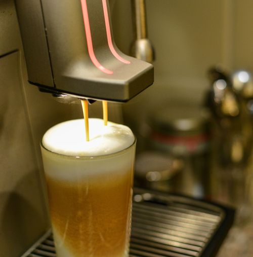 Kaffee läuft aus Vollautomat in ein Glas mit Milchschaum.