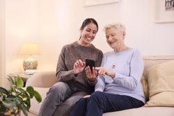 Eine junge Frau und eine ältere Dame sitzen gemeinsam auf einem Sofa. Die jüngere Frau erklärt den Malteser Hausnotruf.