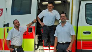 Drei Mitarbeiter des Malteser Rettungsdienstes stehen in Einsatzkleidung am Rettungswagen.