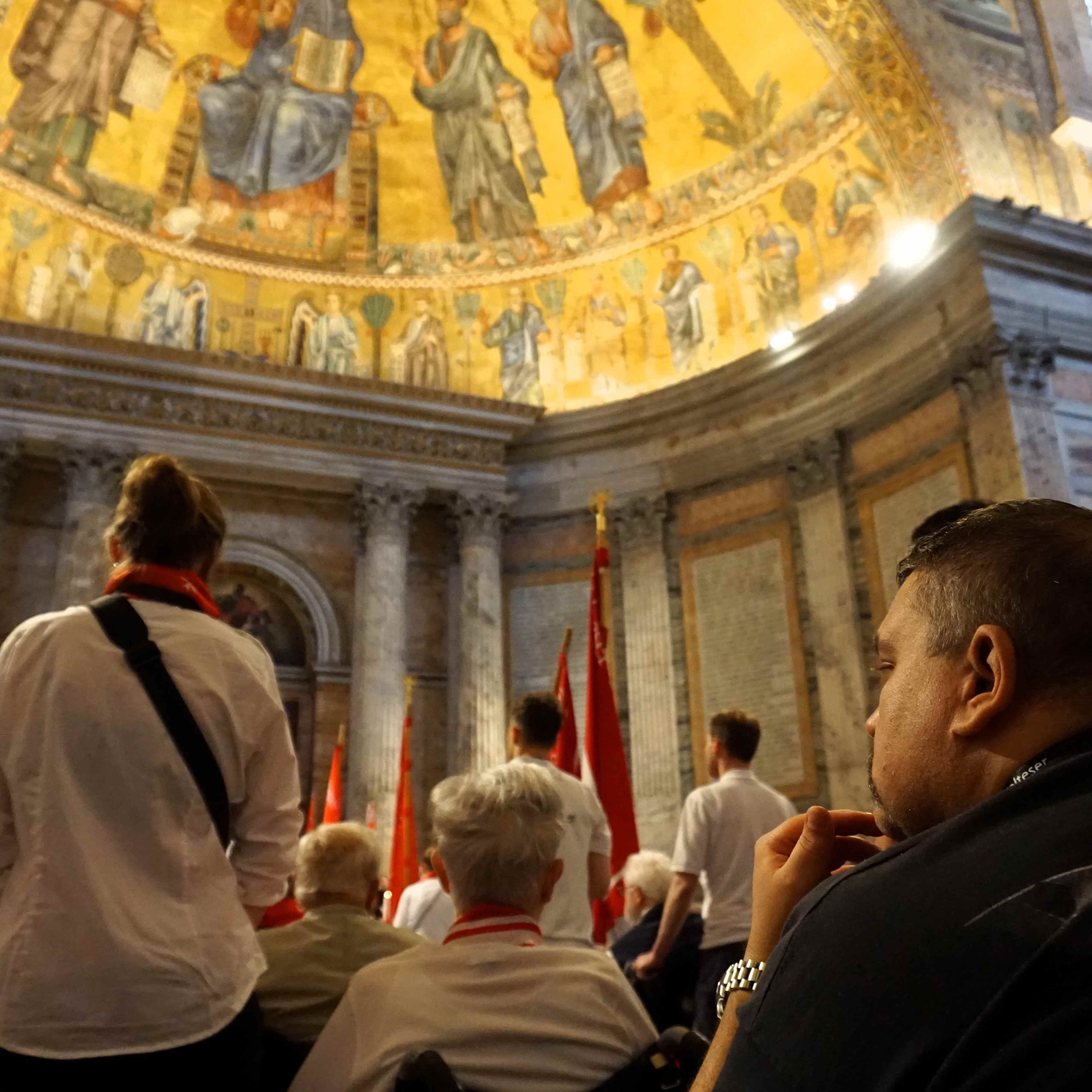 Menschen in einer Kirche mit goldgelber Kuppeldecke und Wandmalereien, einige halten rote Fahnen.