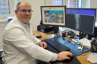 Arzt im weißen Kittel sitzt am Schreibtisch und betrachtet Röntgen- und 3D-Bilder auf zwei Monitoren.