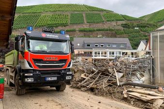 Ein Einsatzfahrzeug des Malteser Hilfsdienstes steht vor einem Haufen Hochwasserschutt in einem Wohngebiet; im Hintergrund sind Weinberge an einem Hang zu sehen. (Foto: Malteser)