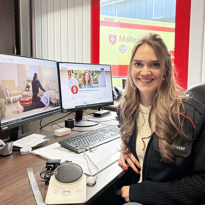 Lächelnde Frau in Malteser Dienstkleidung sitzt vor ihrem Computer.