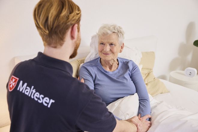 Ein Malteser-Mitarbeiter spricht mit einer älteren Frau im Bett und hält ihre Hand.