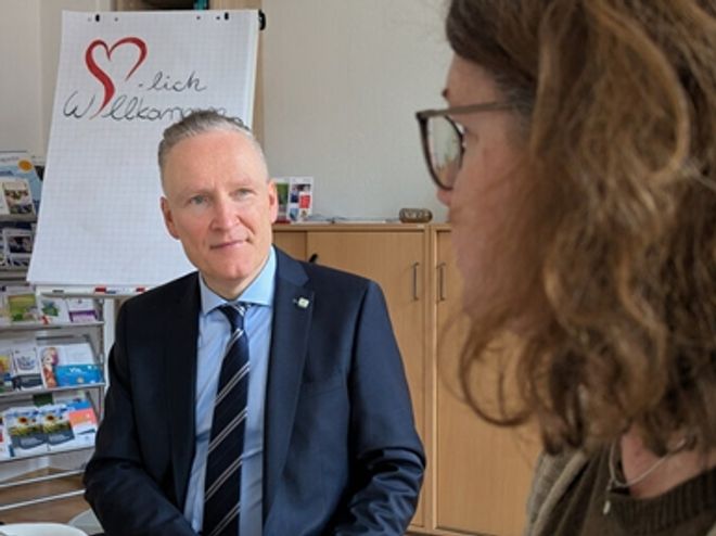 Eine Person in formeller Kleidung sitzt an einem Tisch in einem Innenraum. Im Hintergrund steht ein Flipchart mit der Aufschrift „Herzlich Willkommen“ und einem roten Herzsymbol. Rechts im Bild ist eine weitere Person im Vordergrund zu sehen. Im Raum befinden sich außerdem Informationsmaterialien in einem Regal.