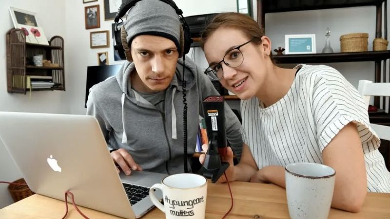 Ein junger Mann und eine junge Frau vor einem Mikrofon und einem Laptop