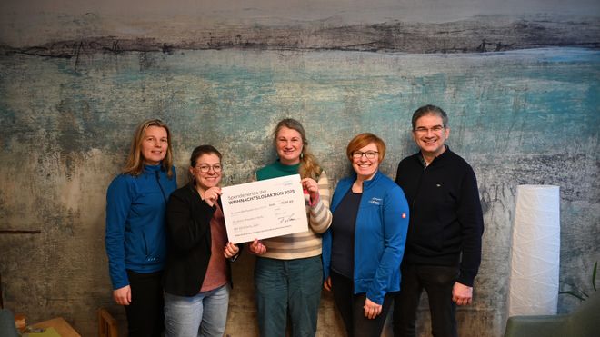 Fünf Personen stehen in einem Raum vor einer blau-grau strukturierten Wand und halten gemeinsam einen großen Spendenscheck. Zwei der Personen tragen blaue Jacken mit Malteser-Aufdruck. Auf dem Scheck steht „Spendenübergabe der Weihnachtsaktion 2023“ sowie die übergebene Summe und Unterschriften. Die Gruppe posiert für ein gemeinsames Foto der Spendenübergabe.