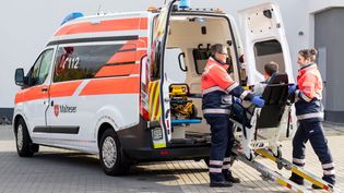Malteser in Einsatzkleidung helfen einem gehbehindertem Mann in in ein Fahrzeug.