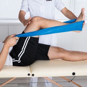 Physiotherapeutin unterstützt liegenden Patienten bei Knieübung mit blauem Fitnessband.
