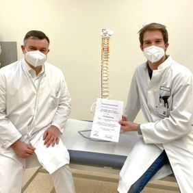 Zwei Ärzte im weißen Kittel sitzen neben einem Wirbelsäulenmodell und halten ein Zertifikat.