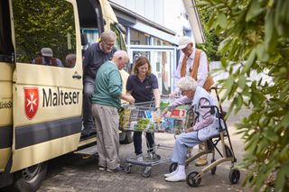 Malteser Mitarbeiterin räumt mit älteren Menschen vor einem Malteser Auto den Einkaufswagen aus