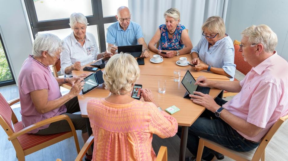 Ältere Menschen mit Kaffeetassen, Tablets und Smartphones an einem Tisch.