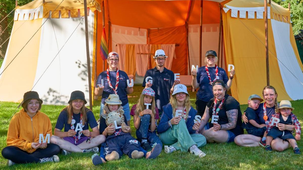 Eine Gruppe von Personen unterschiedlichen Alters sitzt auf einer Wiese vor einem orangenen Zelt.