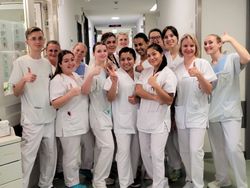 Pflegekräfte in weißer Kleidung stehen lächelnd im Flur eines Krankenhauses und zeigen Daumen hoch.