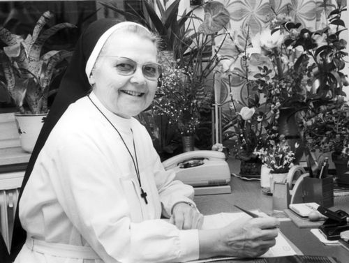 Ordensschwester mit Schreibtisch, umgeben von Pflanzen und Büromaterial.