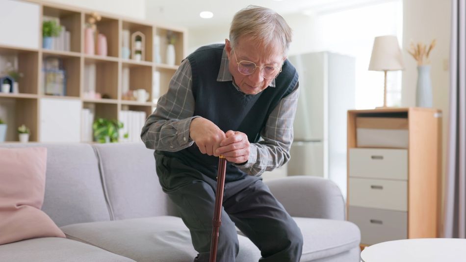 Ein alter Mann mit Gehstock hat Probleme, von einem Sofa aufzustehen.