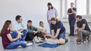 Eine Gruppe von Teilnehmern beobachtet, wie eine Helferin der Malteser die Herzdruckmassage demonstriert