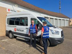 Zwei Ehrenamtliche der Malteser präsentieren die neuen, blauen Warnwesten mit Logo des Mobilen Einkaufswagens vor einem weißen Transporter