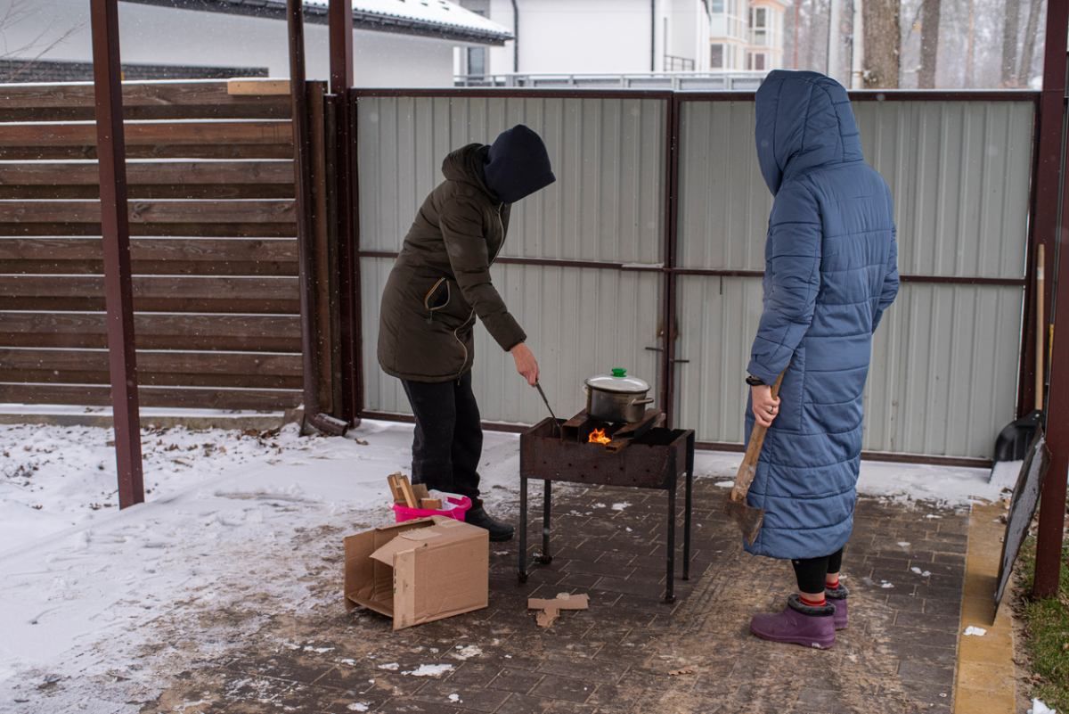 Zwei Menschen in Winterjacken kochen draußen etwas in einem Topf, der auf einem Grill steht.
