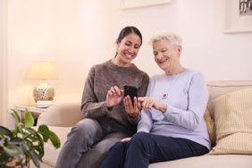 Eine ältere und eine jüngere Frau sitzen auf einem Sofa und schauen gemeinsam auf ein Smartphone.