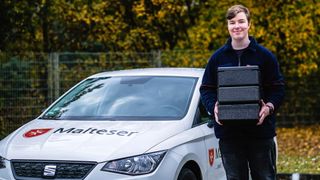 Ein junger Mann trägt Warmhalteboxen und steht vor einem Malteser Fahrzeug. 