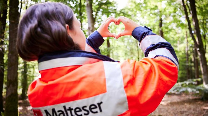 Eine lächelnde Frau mit Spaten und Säcken bei der Landschaftspflege zwischen Bäumen und Gräsern. 