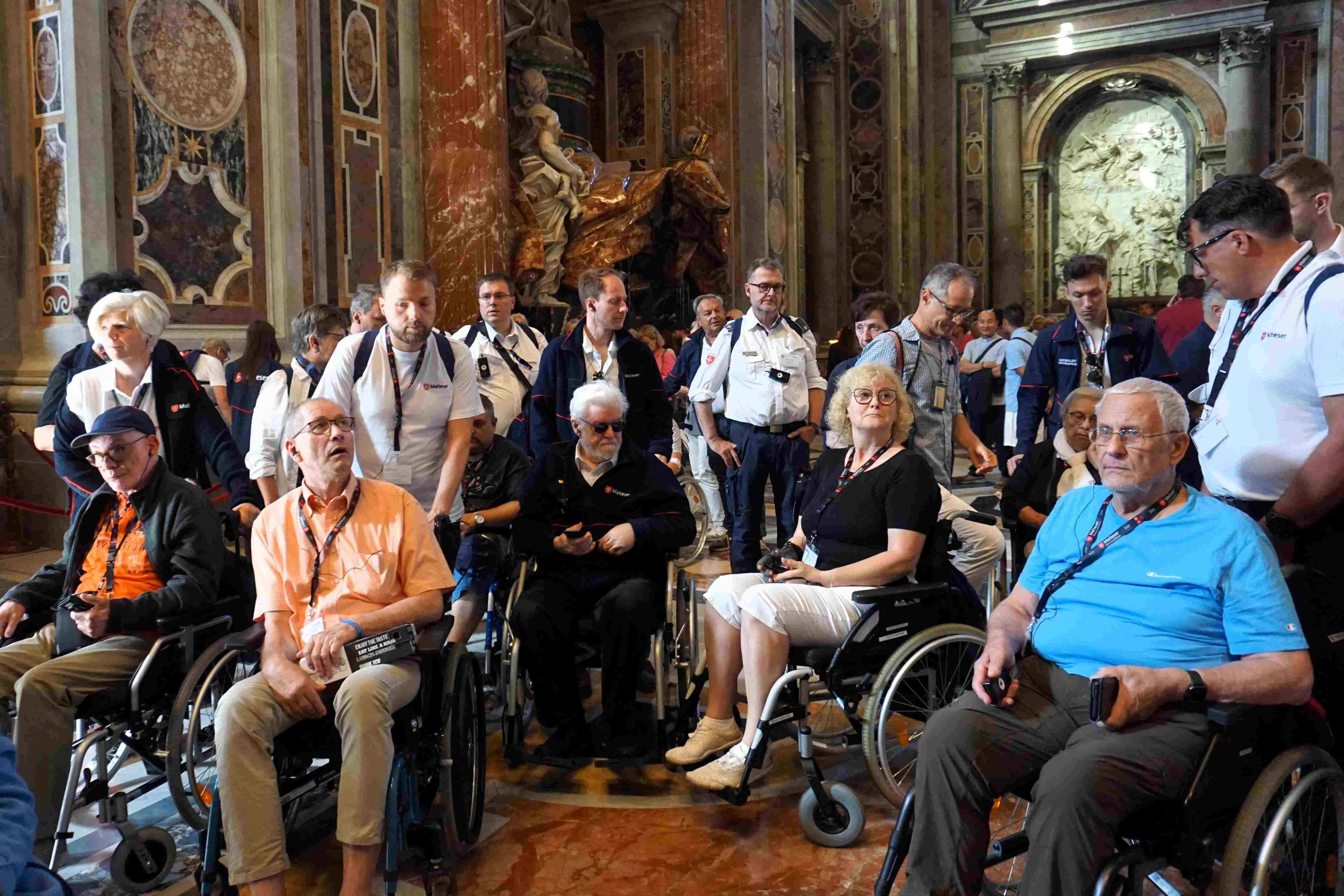 Gruppe von Menschen, einige im Rollstuhl, in einem prunkvoll verzierten Raum mit hohen Decken und großen Türen.