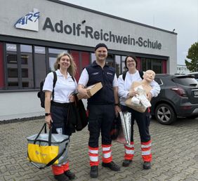 Zwei Sanitäterinnen und ein Sanitäter stehen vor der Limburger Adolf-Reichwein-Schule mit Material für den Wiederbelebungsunterricht. 