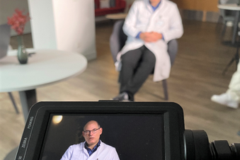Prof. Dr. Tischer in weißem Kittel bei einem Videodreh, sitzend, im Kameradisplay deutlich im Portrait erkennbar.