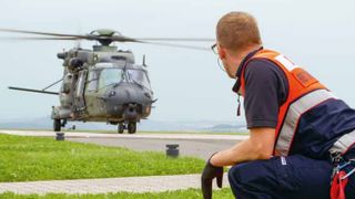 Malteser Helfer hockt auf dem Boden und schaut auf einen landenden Hubschrauber.