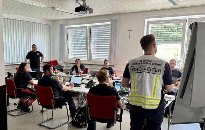 Ausbildungsraum, in dem die Übung stattfindet. Die Teilnehmenden sitzen an Tischen, der Übungsleiter steht mit dem Rücken zur Kamera.