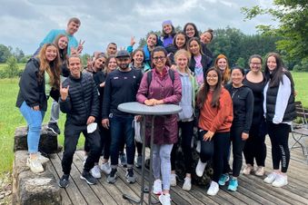 Gruppe junger Menschen posiert fröhlich im Grünen auf Holzterrasse vor bewölktem Himmel – gemeinsamer Ausflug oder Team-Event.