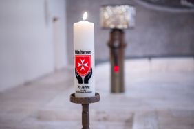 Eine brennende Kerze mit Malteser-Logo steht in einer Kirche vor einem Altar.