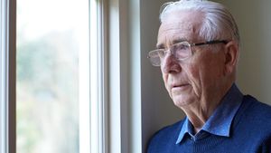 Ein älterer Mann mit Brille und blauem Pullover schaut besorgt aus dem Fenster.