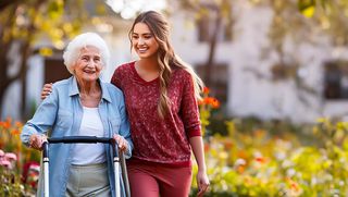 Eine jüngere Frau geht mit einer älteren Frau spazieren, die einen Rollator nutzt. 