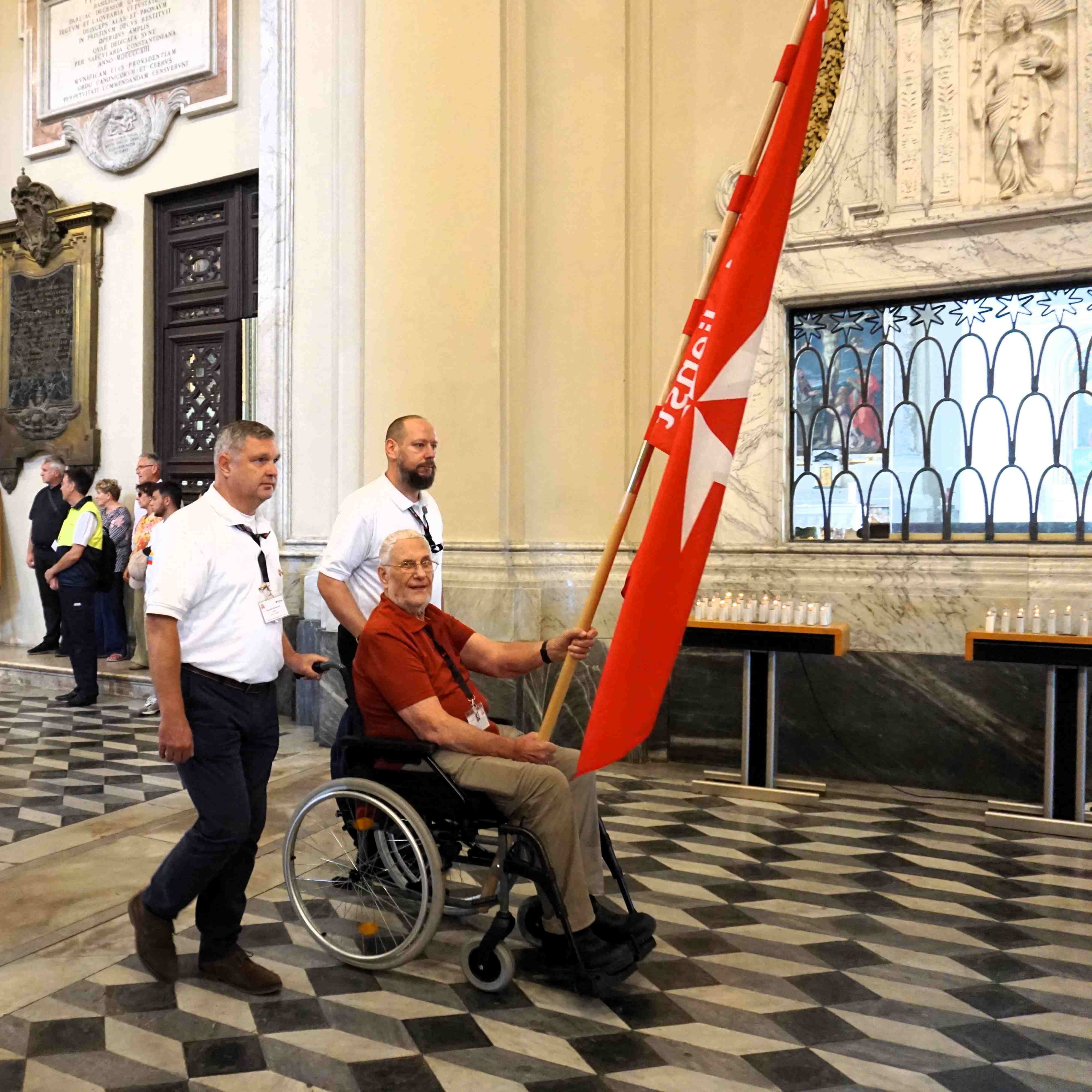 Mann im Rollstuhl hält rote Bannerfahne mit weißem Kreuz, begleitet von zwei Männern in weißem Hemd in einem historischen Innenraum mit schwarz-weißem Schachbrettmuster auf dem Boden.