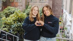 Zwei junge Frauen im Malteser Pullover lächeln in die Kamera und zeigen mit den Händen ein Herz.