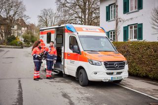 Ein Rettungswagen parkt vor einem Haus. Davor stehen eine Rettungssanitäterin und ein Rettungssanitäter. 